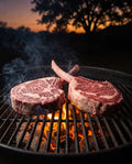 Two Hassell Wagyu Gold Tomahawk steaks on a charcoal grill grate at dusk