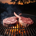 Two Hassell Wagyu Gold Tomahawk steaks on a charcoal grill grate at dusk